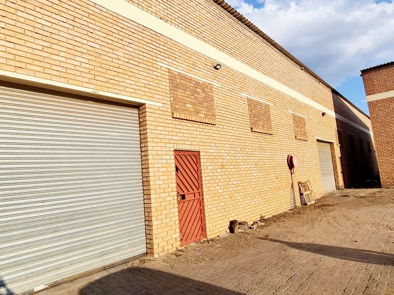 Exterior view of an industrial warehouse facility featuring brick construction, large roller shutter doors, and a paved yard area.