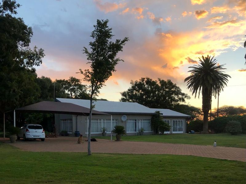 A charming farmhouse exterior featuring a paved driveway, a covered carport, and a spacious lawn set against a scenic sunset backdrop.