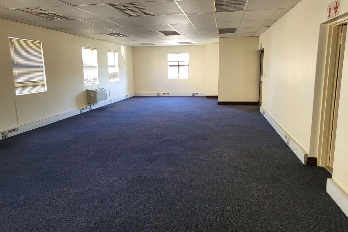 A spacious, empty open-plan office area featuring a suspended ceiling, blue carpeting, and multiple windows providing natural light.