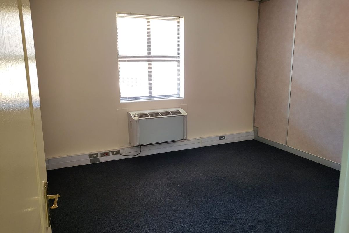 A vacant office space featuring dark carpeting, a wall-mounted air conditioning unit, and neutral wall finishes with integrated electrical trunking.