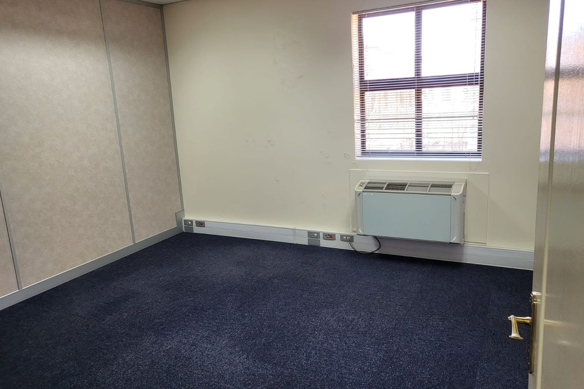 A vacant office space featuring dark carpeting, a wall-mounted air conditioning unit, and a window with fitted blinds.