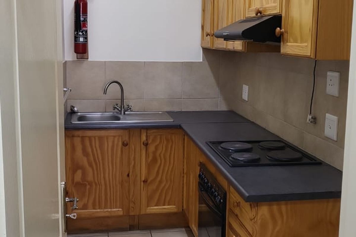 A compact office kitchenette featuring wooden cabinetry, a built-in electric hob, oven, extractor fan, and a stainless steel sink with tiled splashbacks.