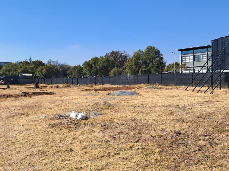 A large, undeveloped plot of land featuring dry grass, perimeter fencing, and a large billboard structure, situated in an office park environment.