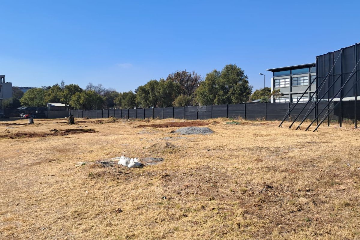 A large, undeveloped plot of land featuring dry grass, perimeter fencing, and a large billboard structure, situated in an office park environment.
