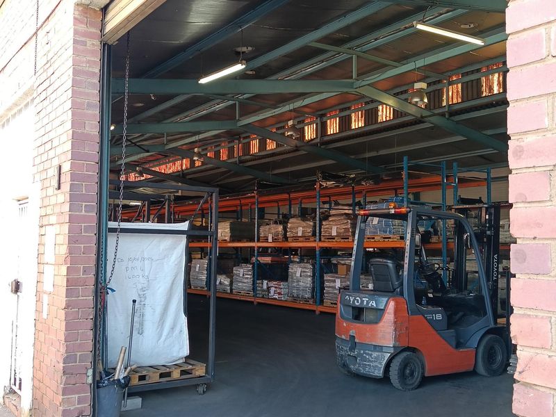 Industrial warehouse space featuring exposed steel roof trusses, high-capacity storage racking, and concrete flooring, accessible via a large roller door entrance.