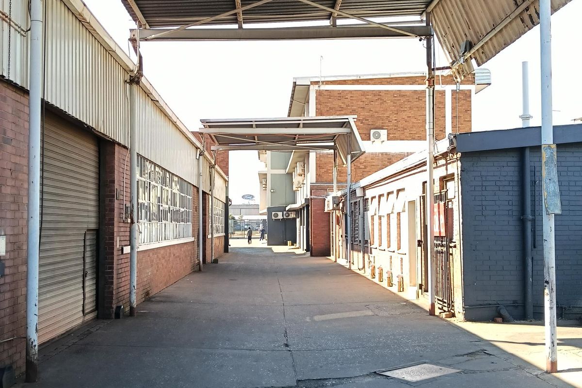 Industrial exterior corridor featuring a covered loading area, concrete driveway, and brick building facades with roller shutter access.