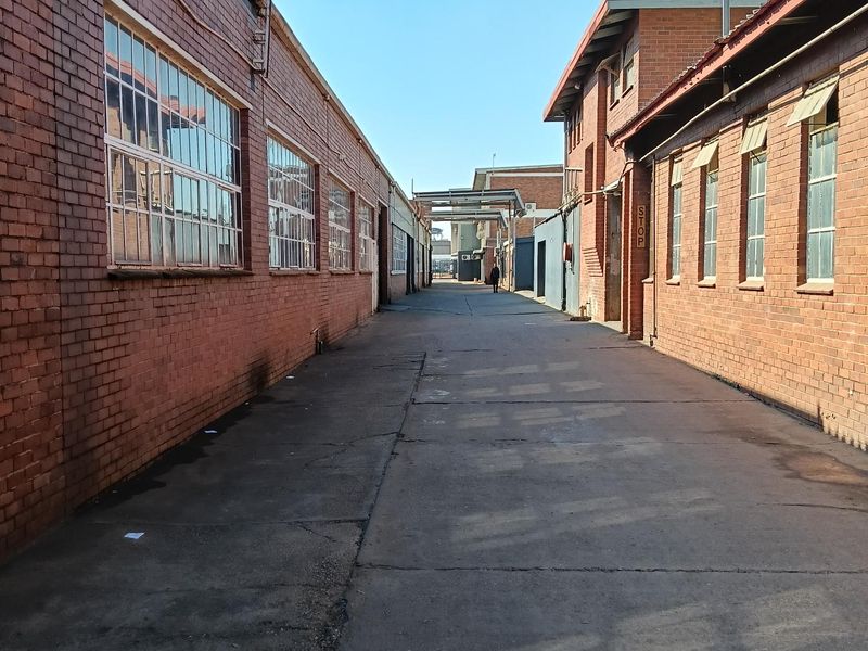 An industrial exterior alleyway featuring exposed brick facades, steel-framed windows, and a concrete driveway connecting the warehouse structures.
