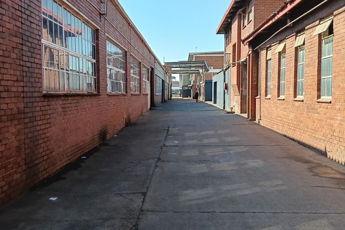 An industrial exterior alleyway featuring exposed brick facades, steel-framed windows, and a concrete driveway connecting the warehouse structures.