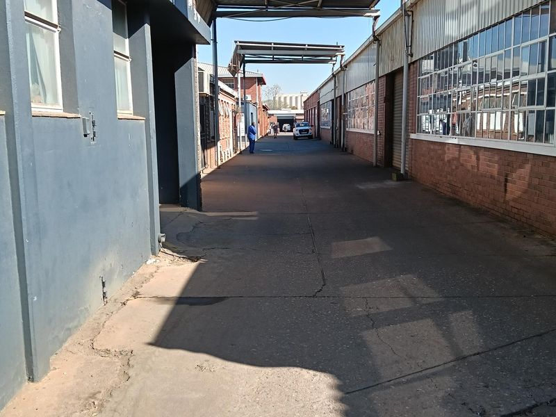 An industrial exterior corridor featuring concrete paving, steel-framed windows, and overhead steel canopy structures between two warehouse buildings.