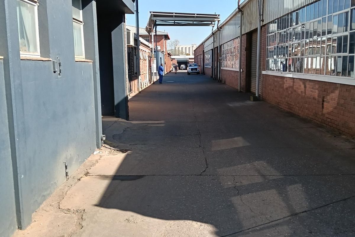 An industrial exterior corridor featuring concrete paving, steel-framed windows, and overhead steel canopy structures between two warehouse buildings.