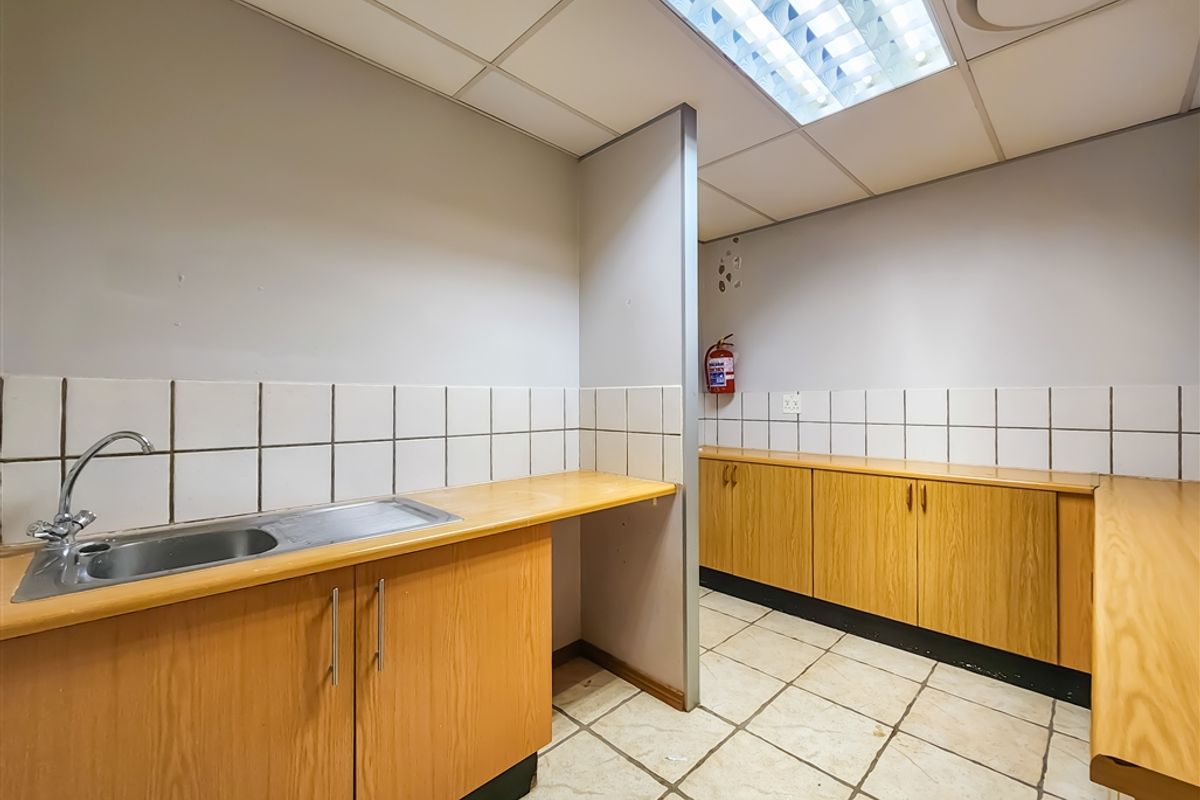 A functional office kitchenette featuring wood-look cabinetry, tiled splashbacks, a stainless steel sink, and recessed fluorescent lighting.