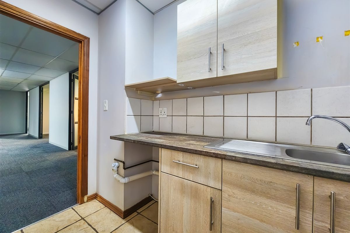 A functional office kitchenette featuring light wood-finish cabinetry, a stainless steel sink, tiled backsplash, and tiled flooring, with direct access to the main office area.