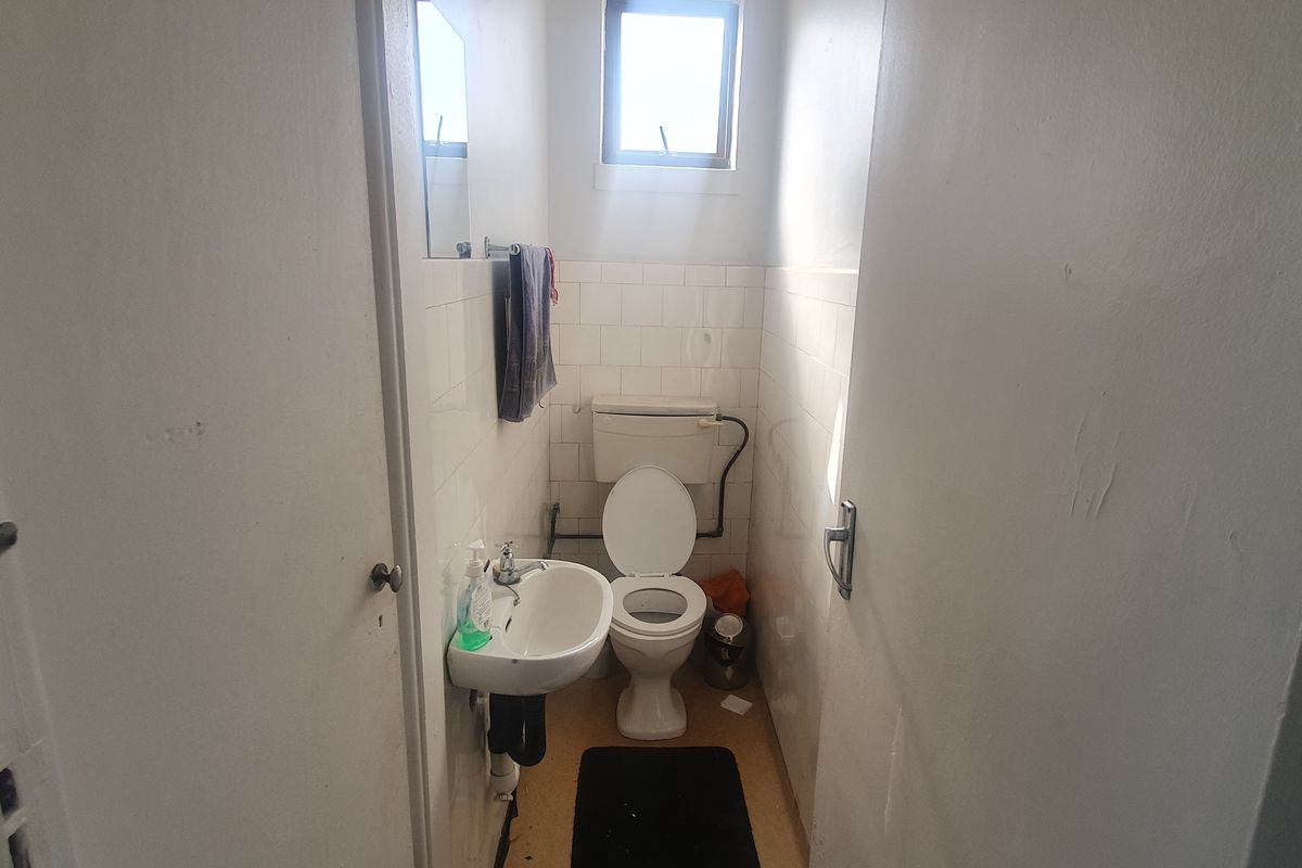 A compact office bathroom featuring a wall-mounted basin, toilet, and tiled splashback, requiring cosmetic updates to improve its dated appearance.