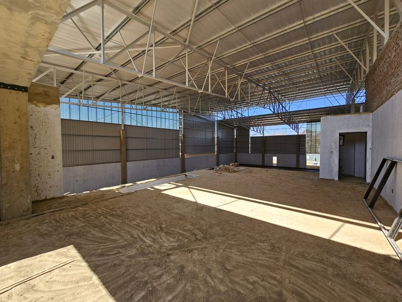 Large industrial utility space featuring exposed steel roof trusses, corrugated metal cladding, and unfinished concrete flooring, currently in a shell construction state.