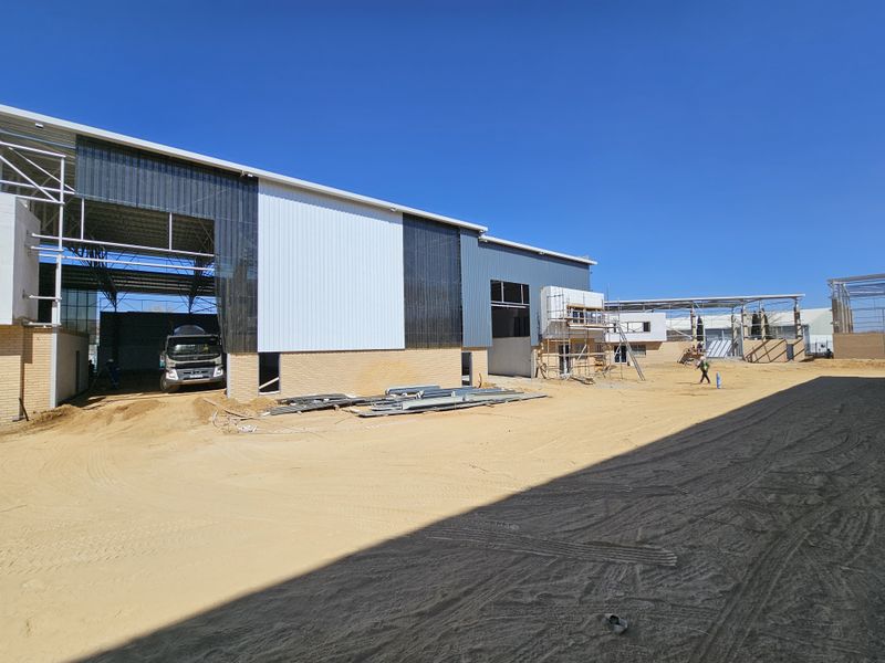 View of an industrial property under construction, featuring steel portal frame structures, metal cladding, and brickwork base on an unpaved site.