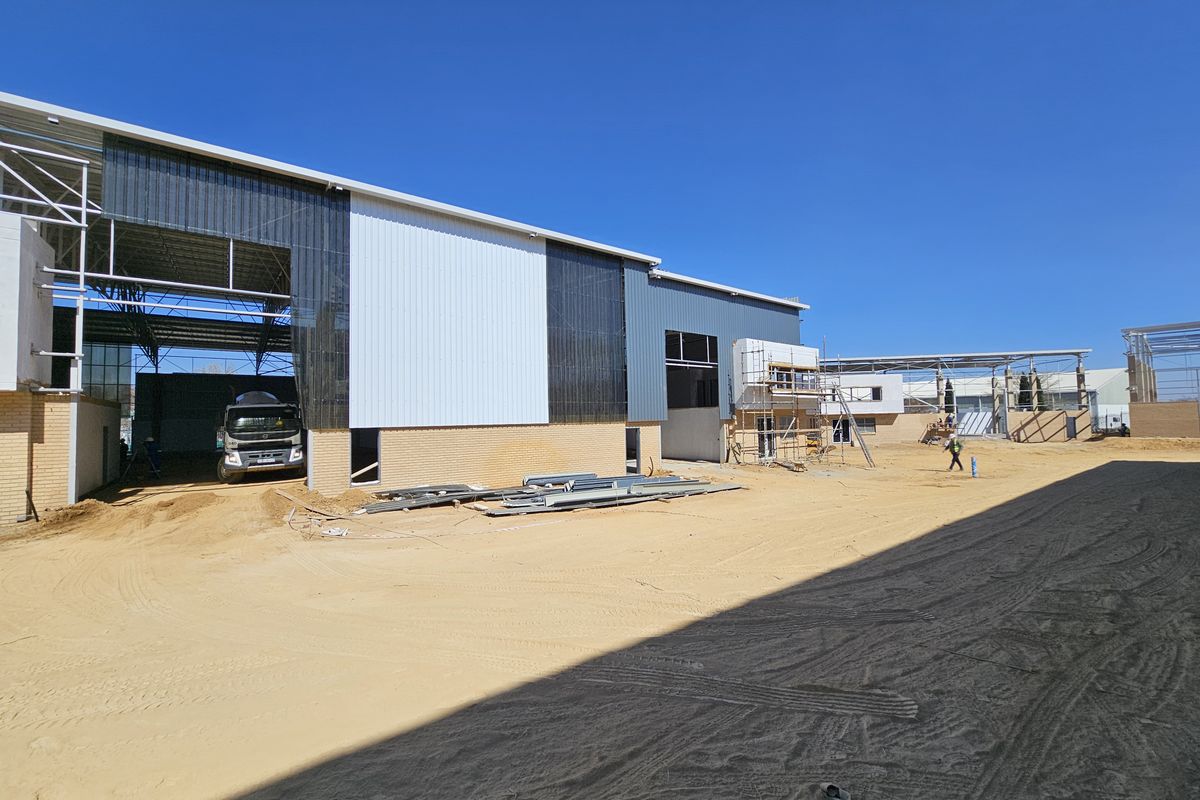 View of an industrial property under construction, featuring steel portal frame structures, metal cladding, and brickwork base on an unpaved site.