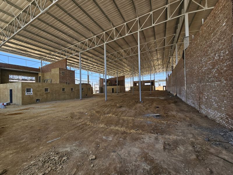 Large industrial warehouse space under construction featuring a steel portal frame, exposed brick walls, and an unfinished dirt floor.
