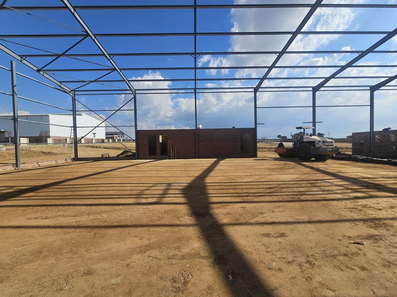 An industrial construction site featuring an exposed steel portal frame structure and partial brick masonry walls on an unpaved earth foundation.