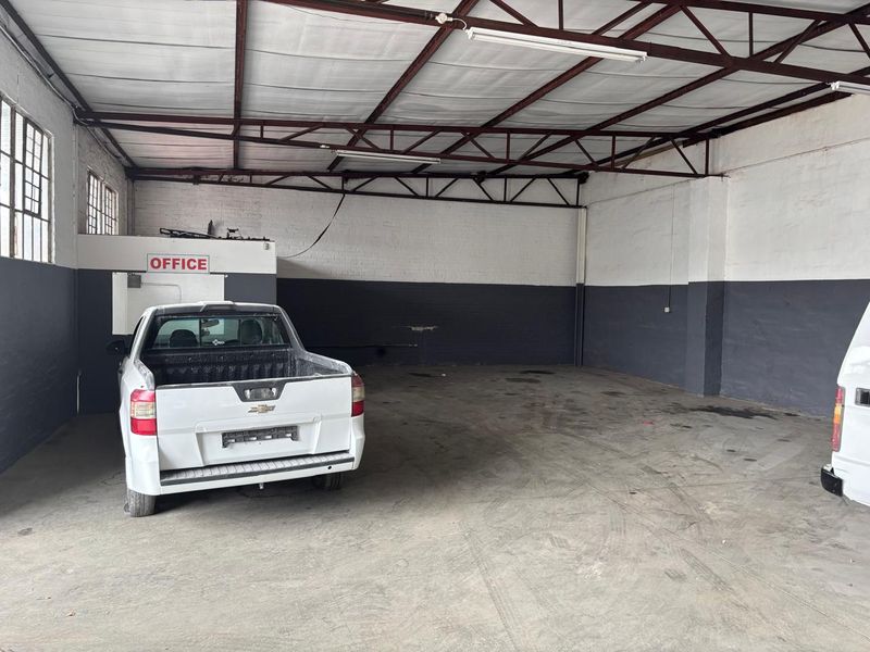Spacious industrial garage or warehouse area featuring exposed steel roof trusses, concrete flooring, and a dedicated enclosed office space.