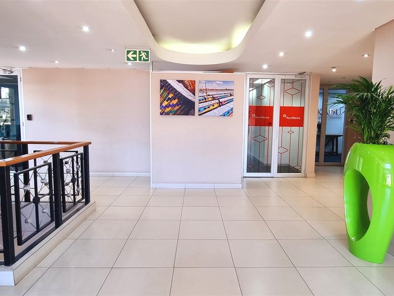 A bright and spacious office building entrance lobby featuring tiled flooring, recessed ceiling lighting, and a modern metal balustrade overlooking the stairwell.