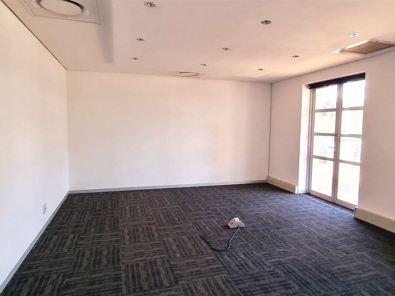 A spacious, empty office area featuring dark carpet tiles, recessed ceiling lighting, and natural light from large windows and a glass door.