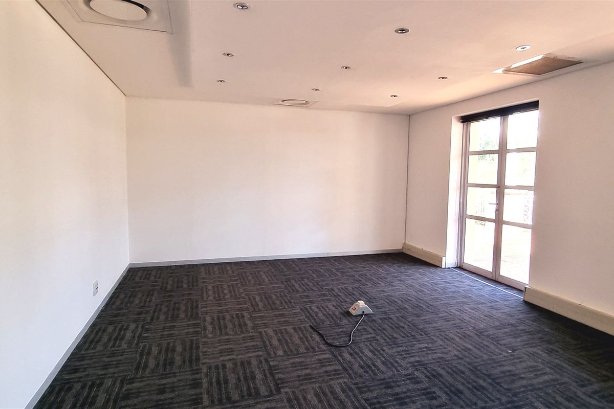 A spacious, empty office area featuring dark carpet tiles, recessed ceiling lighting, and natural light from large windows and a glass door.