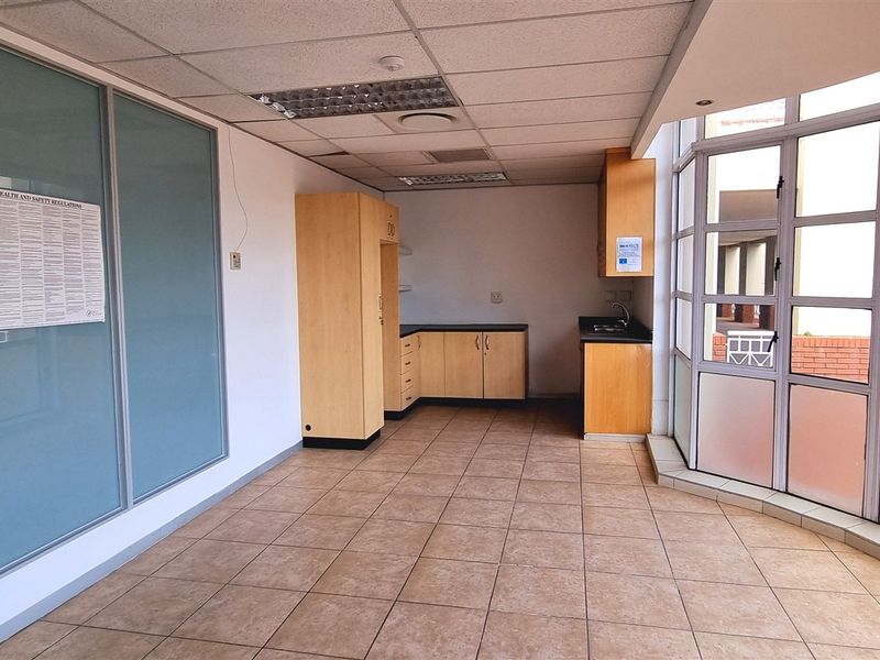 An open-plan office space featuring tiled flooring, a suspended ceiling, a small built-in kitchenette, and large glass windows providing natural light.