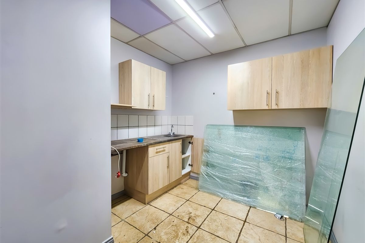 A small office kitchenette featuring light wood-look cabinetry, a stainless steel sink, and tiled flooring, requiring some maintenance and cosmetic updates.
