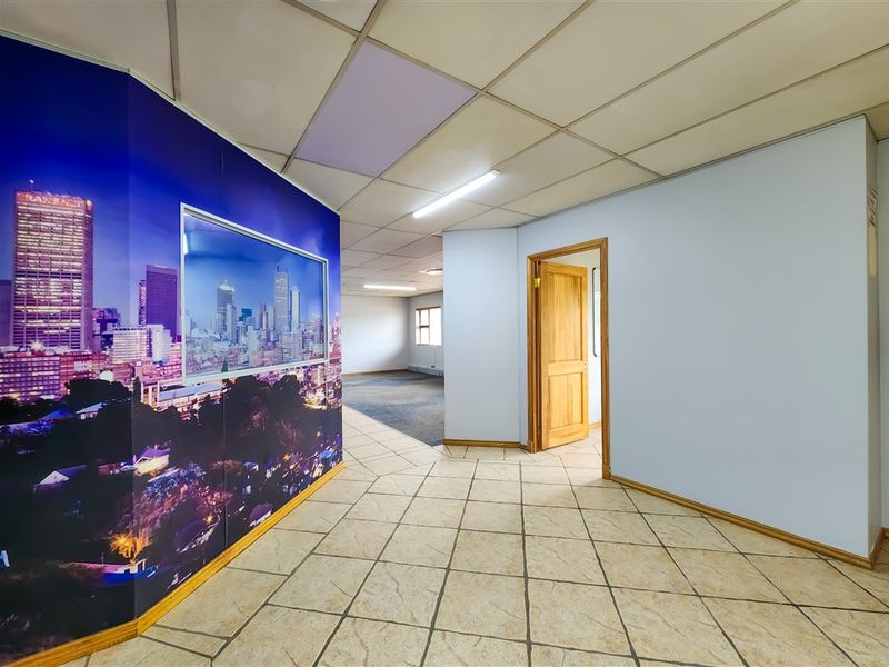 Spacious office interior featuring tiled flooring, suspended ceiling with integrated lighting, and a wooden door leading to an adjacent carpeted workspace.