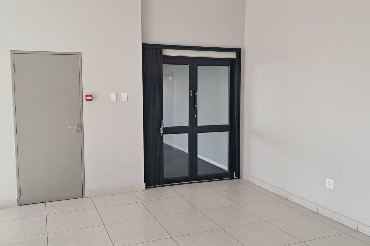 A bright office lobby area featuring tiled flooring, a solid grey door, and a modern black-framed glass double door entrance.