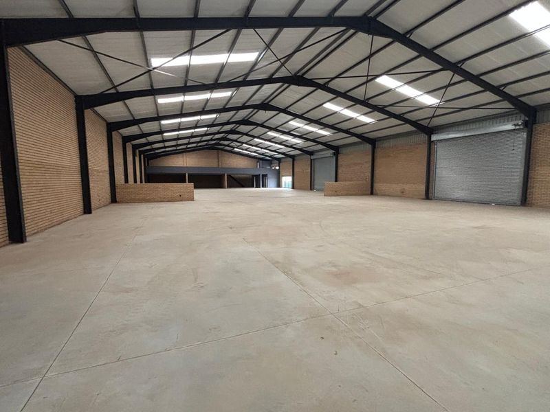 Spacious industrial warehouse featuring high ceilings with exposed steel trusses, concrete flooring, face brick walls, and large roller shutter doors for easy access.