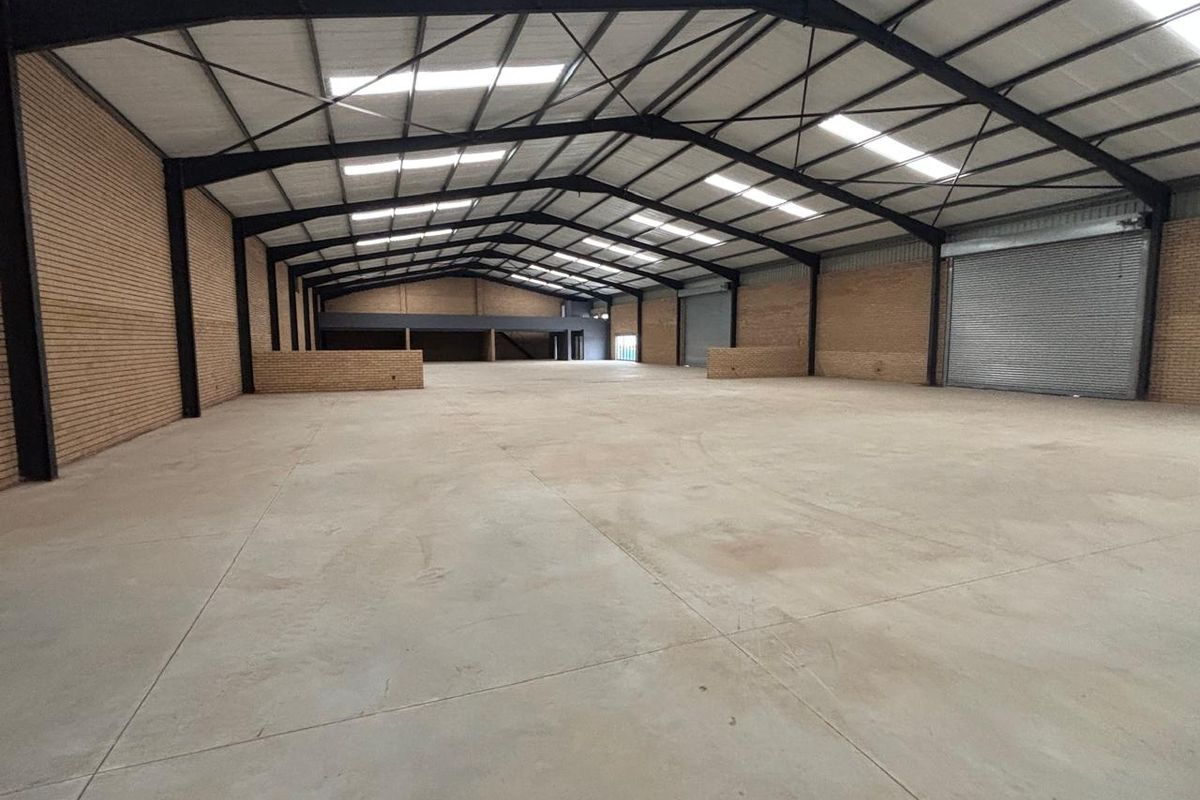 Spacious industrial warehouse featuring high ceilings with exposed steel trusses, concrete flooring, face brick walls, and large roller shutter doors for easy access.