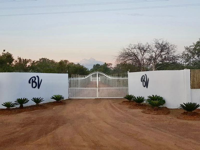 A secure gated entrance to the farm property featuring a white perimeter wall, metal gate, and landscaped garden beds along the driveway.