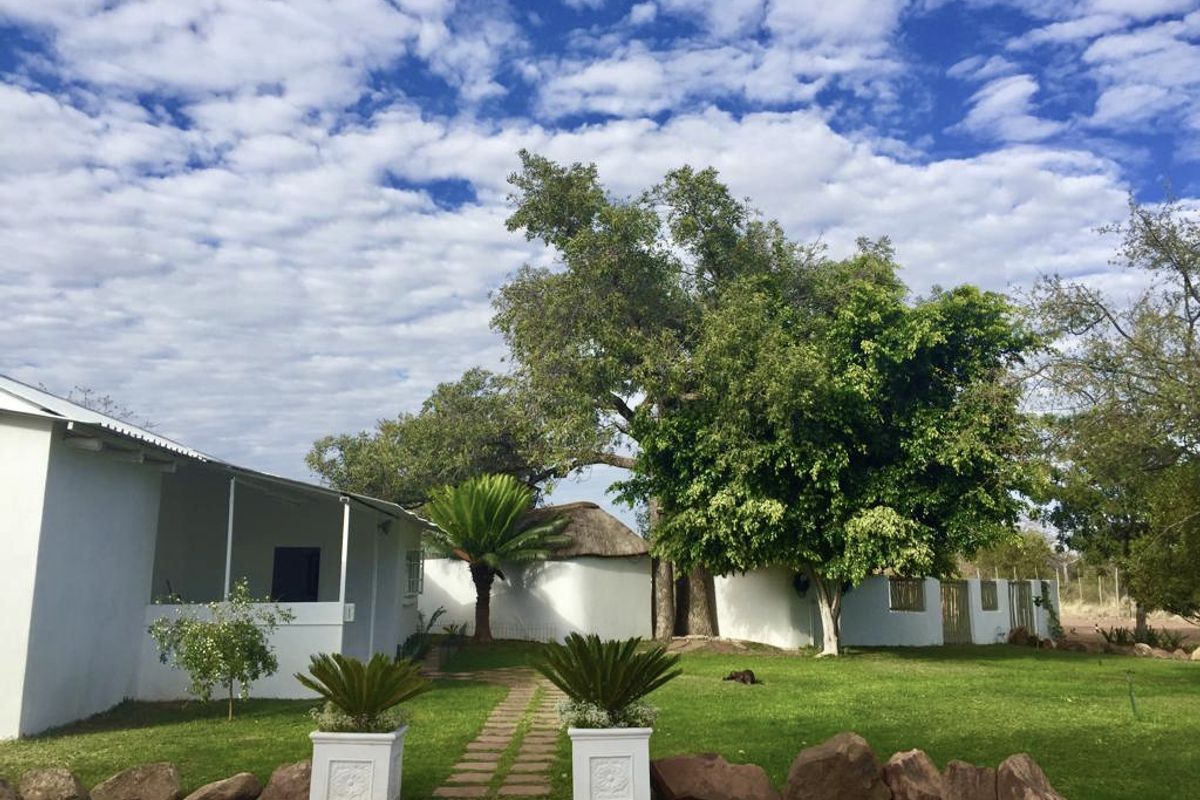 A well-maintained rear garden area featuring a stone pathway, mature trees, a covered patio, and a charming thatched-roof outbuilding on a farm property.