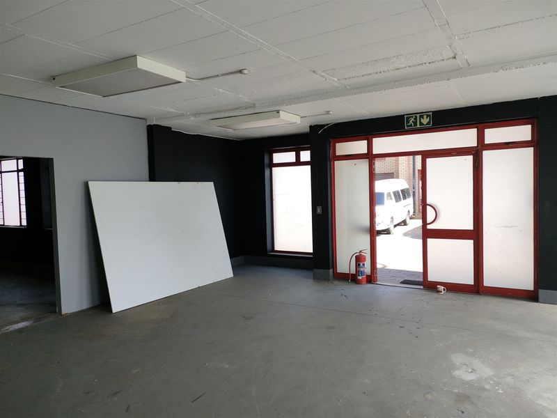 Spacious industrial entry area featuring concrete flooring, fluorescent lighting, and a large aluminium-framed glass door with an emergency exit sign.