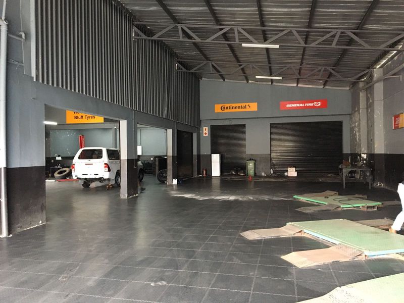 Spacious commercial garage workshop featuring high ceilings with exposed steel trusses, roller shutter doors, and durable tiled flooring suitable for automotive services.