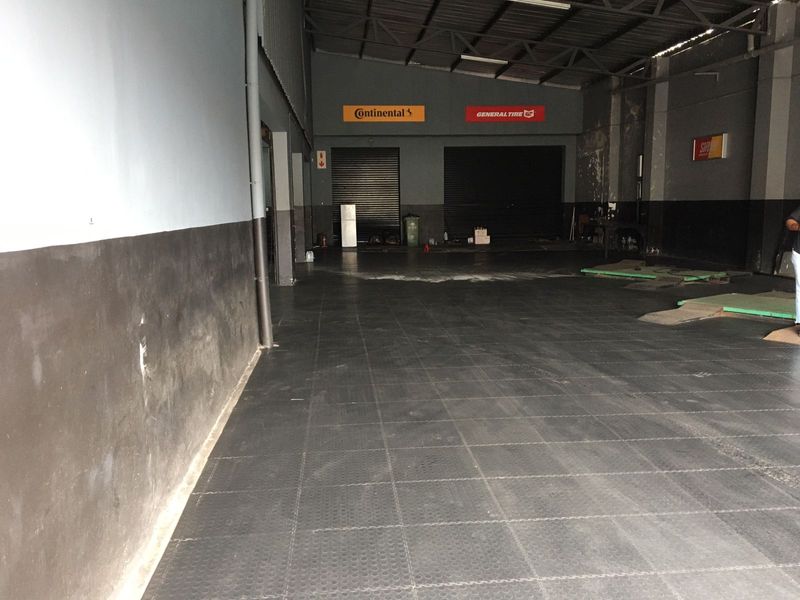 Spacious commercial garage or workshop area featuring high ceilings with exposed steel trusses, roller shutter doors, and durable interlocking floor tiles.