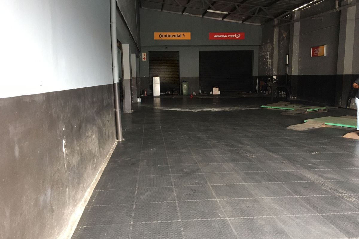 Spacious commercial garage or workshop area featuring high ceilings with exposed steel trusses, roller shutter doors, and durable interlocking floor tiles.