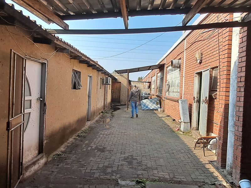 A narrow exterior passage between industrial buildings featuring paved flooring, brick and plastered walls, and a partial corrugated metal roof covering.