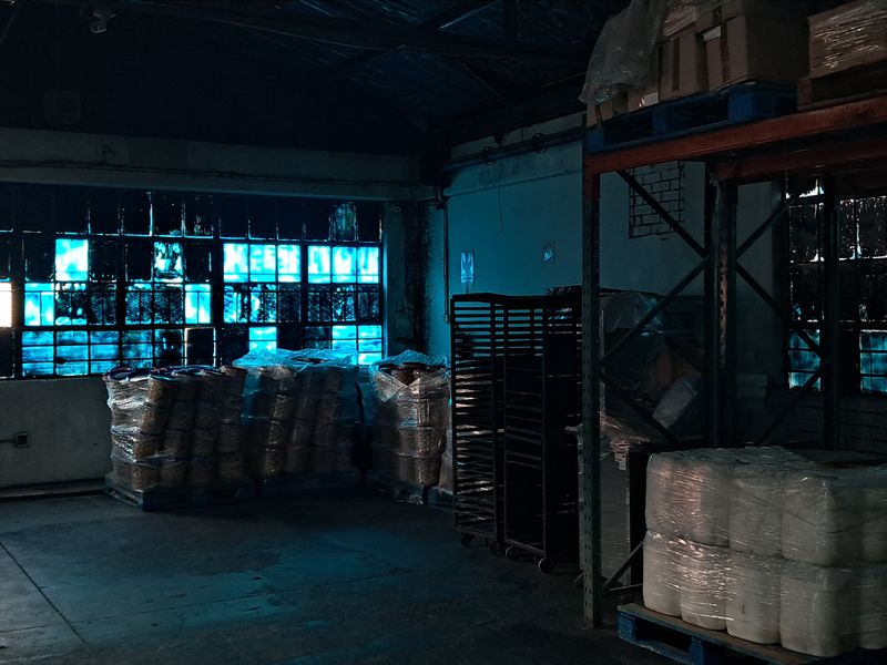 Industrial warehouse space featuring large steel-framed windows, concrete flooring, and exposed structural elements, currently utilized for storage.