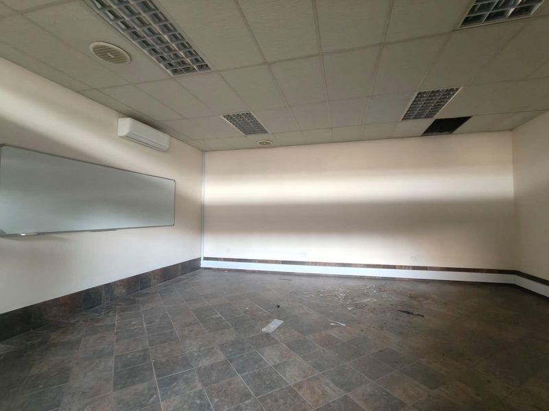 An empty office space featuring tiled flooring, a suspended ceiling with recessed lighting, a wall-mounted air conditioning unit, and a whiteboard.