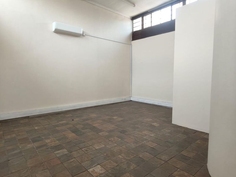 A vacant office space featuring tiled flooring, high ceilings with clerestory windows, and a wall-mounted split-system air conditioning unit.