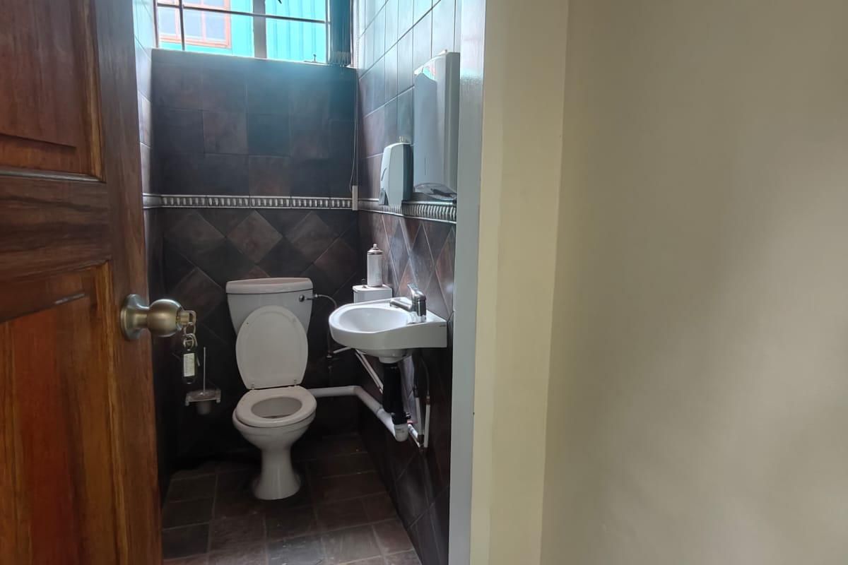 A compact office bathroom featuring dark wall and floor tiling, a wall-mounted basin, and a standard toilet with exposed plumbing.