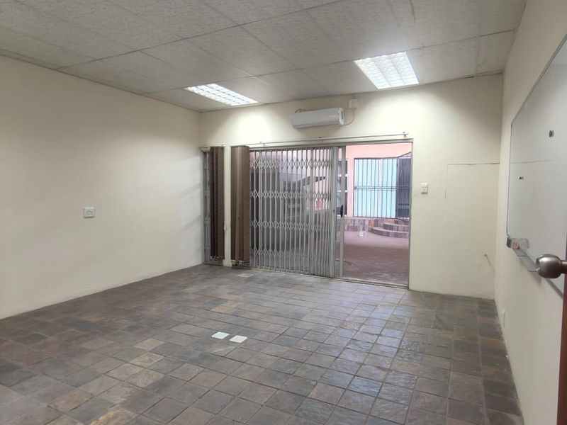 An empty office space featuring tiled flooring, a suspended ceiling with recessed lighting, a wall-mounted air conditioner, and a security gate leading outdoors.