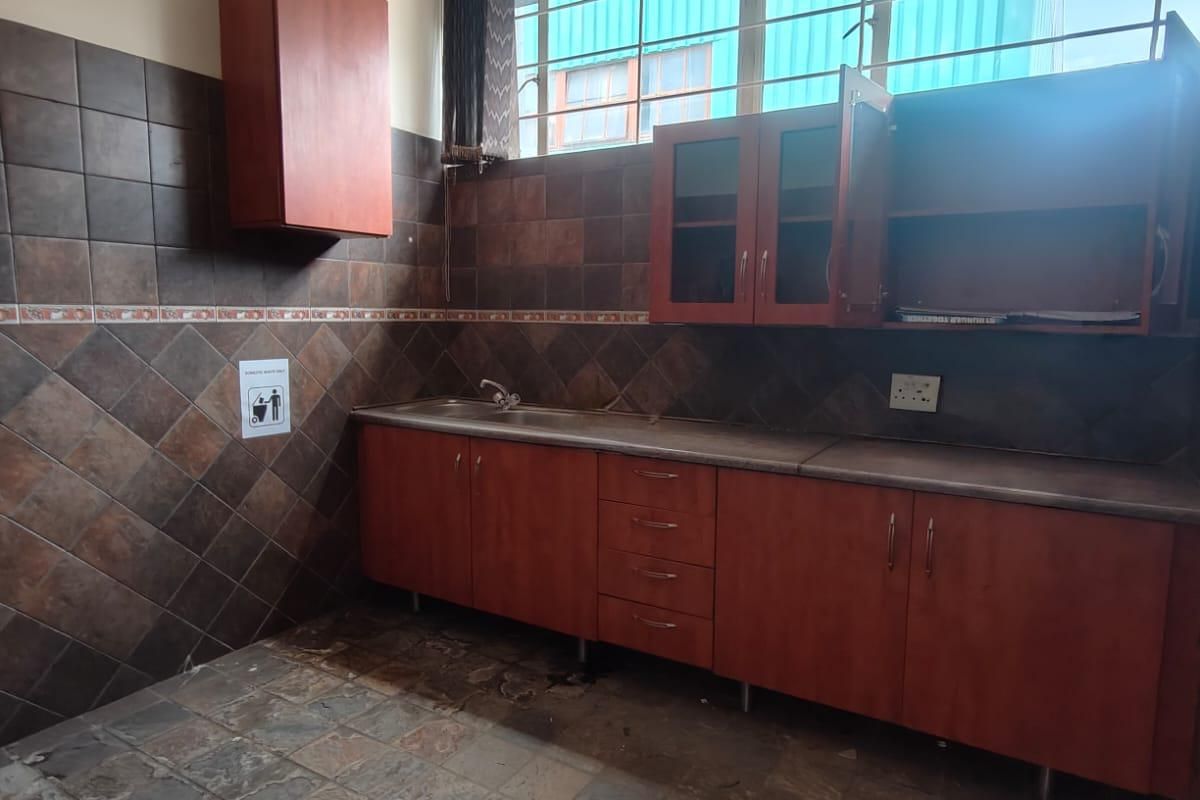 A functional office kitchenette featuring wood-finish cabinetry, a stainless steel sink, tiled backsplash, and durable floor tiles, suitable for staff use.