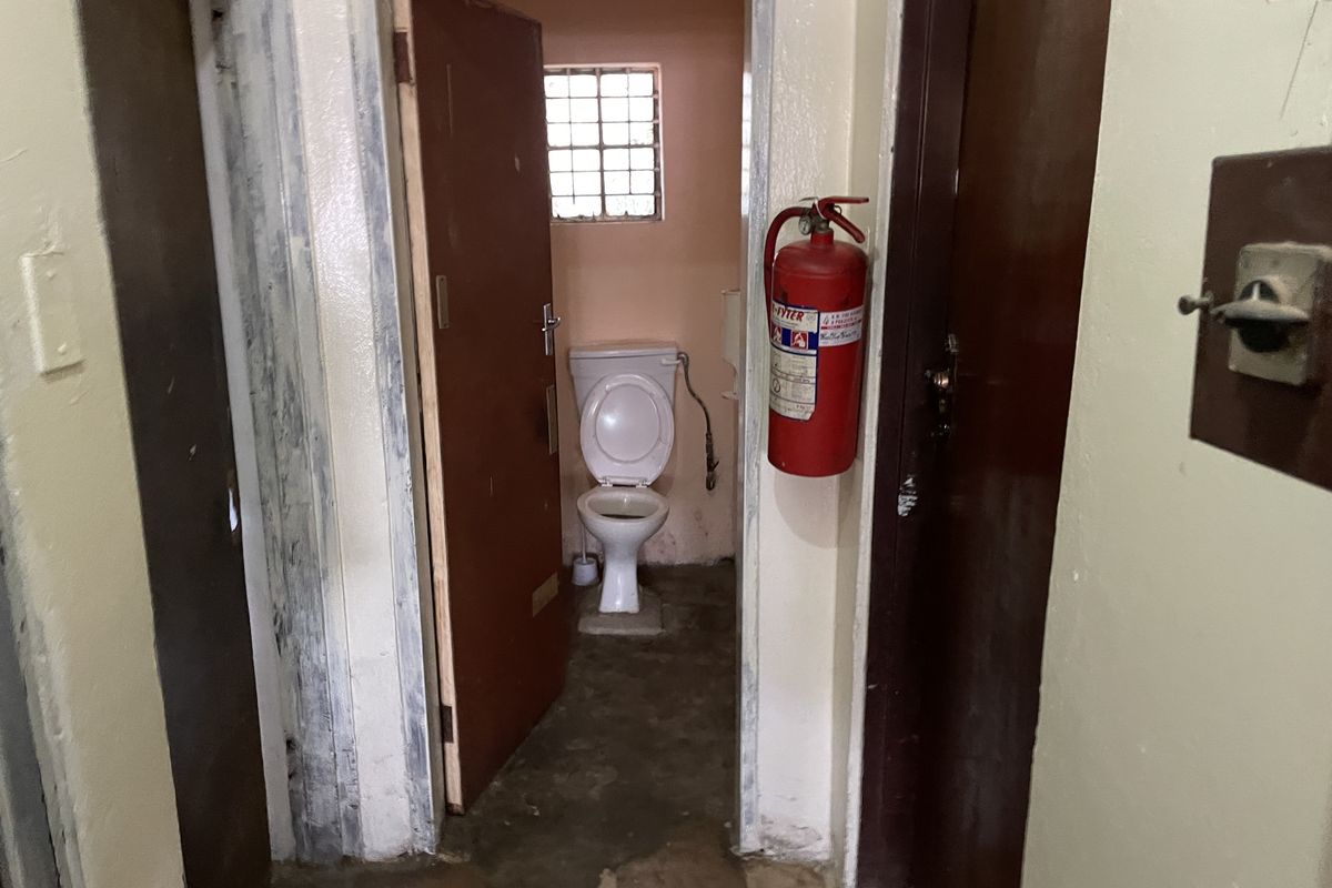 A small, dated bathroom facility featuring a standard toilet, security window grille, and wall-mounted fire extinguisher, requiring significant maintenance and cosmetic updates.
