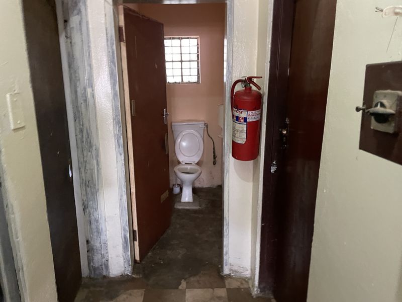 A small, dated bathroom facility featuring a standard toilet, security window grille, and wall-mounted fire extinguisher, requiring significant maintenance and cosmetic updates.