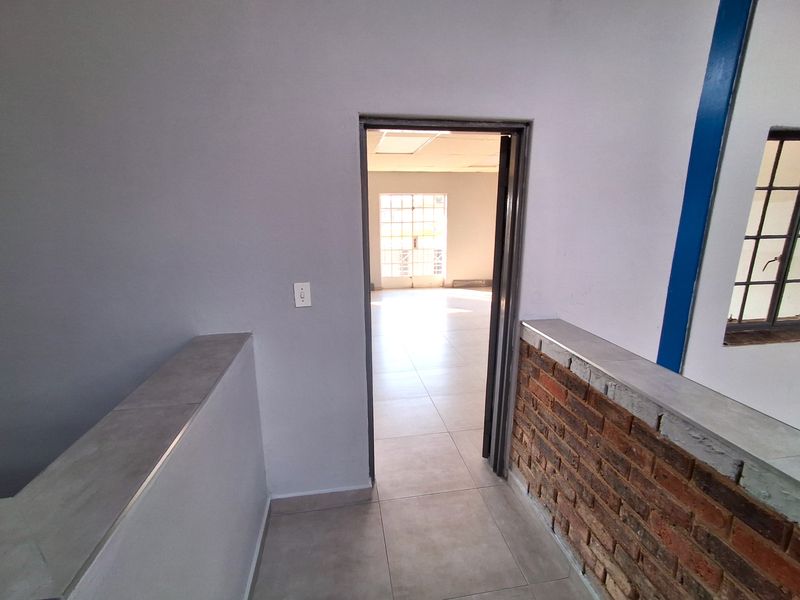 A bright, empty hallway featuring tiled flooring and an exposed brick accent wall, leading into an adjacent open-plan industrial space.