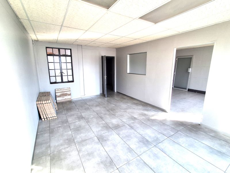 A clean, vacant office space featuring neutral grey tiled flooring, a suspended ceiling, and an internal window providing natural light flow between rooms.