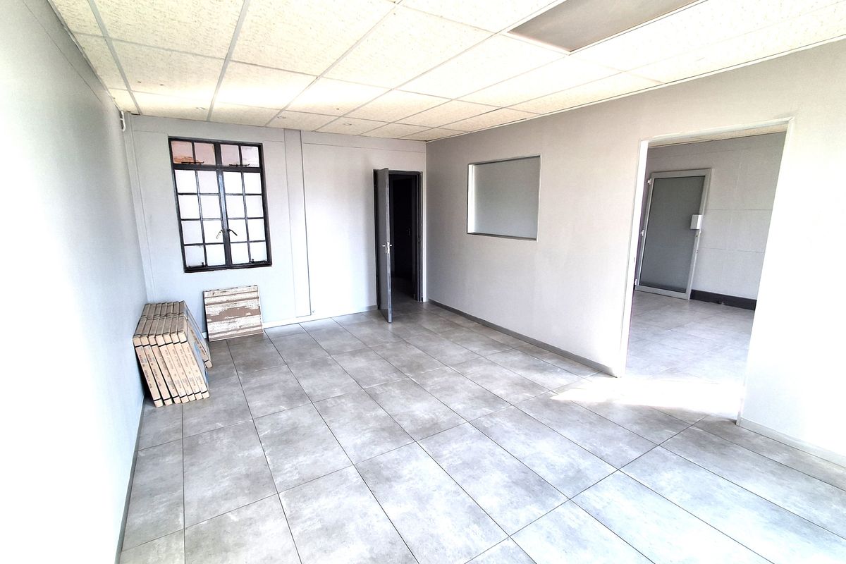 A clean, vacant office space featuring neutral grey tiled flooring, a suspended ceiling, and an internal window providing natural light flow between rooms.